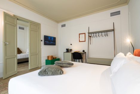 Interno di design della camera Luigi: pavimento in cementine siciliane originali, soffitto decorato, arredi minimalisti e appendiabiti a vista. Sullo sfondo, scorcio della stanza adiacente attraverso le porte d'epoca.