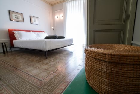 Vista prospettica della Stanza Luigi a Castellett Ragusa: in primo piano un cesto in vimini intrecciato, sullo sfondo il letto matrimoniale con testiera rossa e il pavimento d'epoca decorato.
